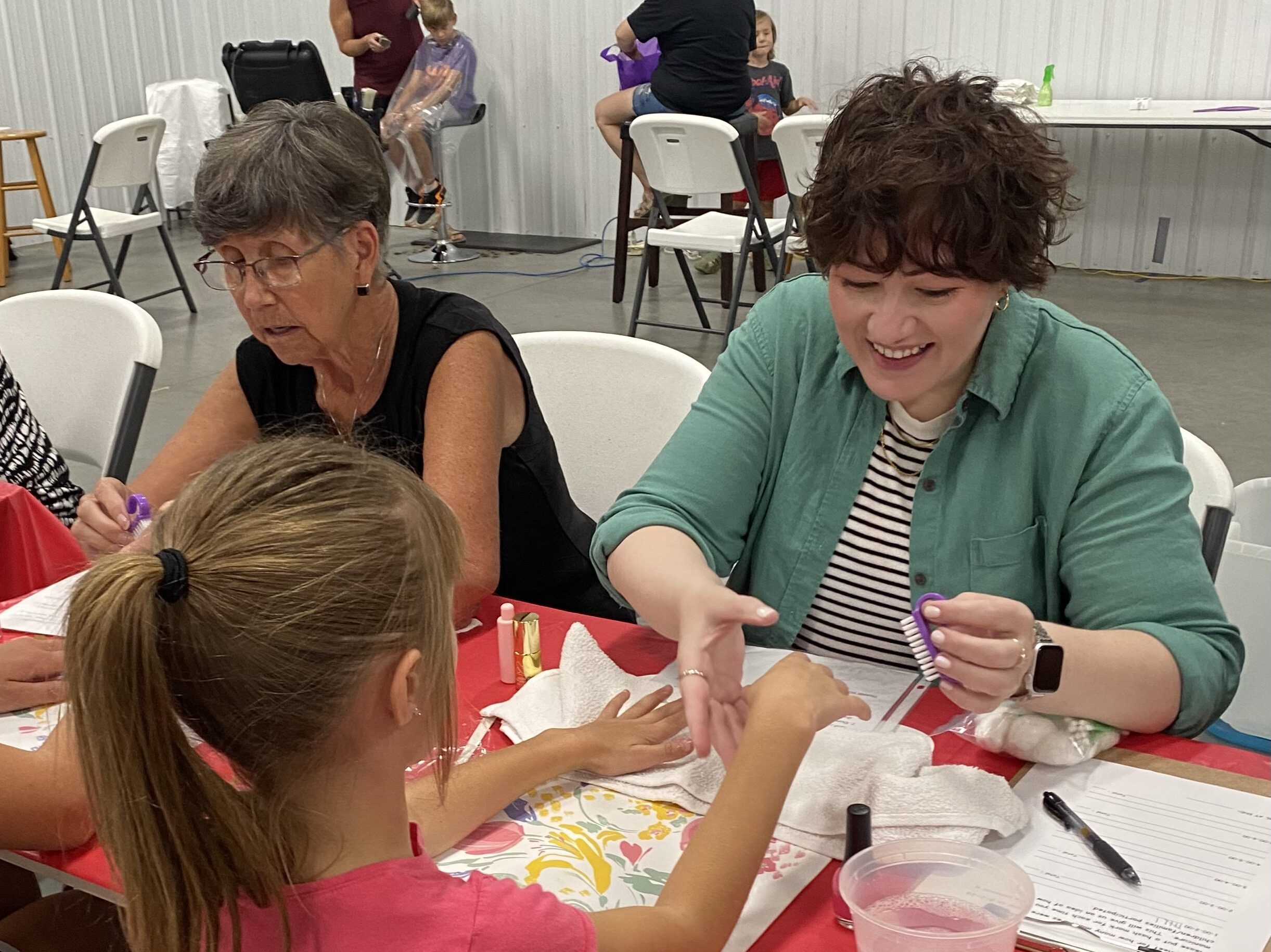 A woman smiles as she paints a young girls fingernails.