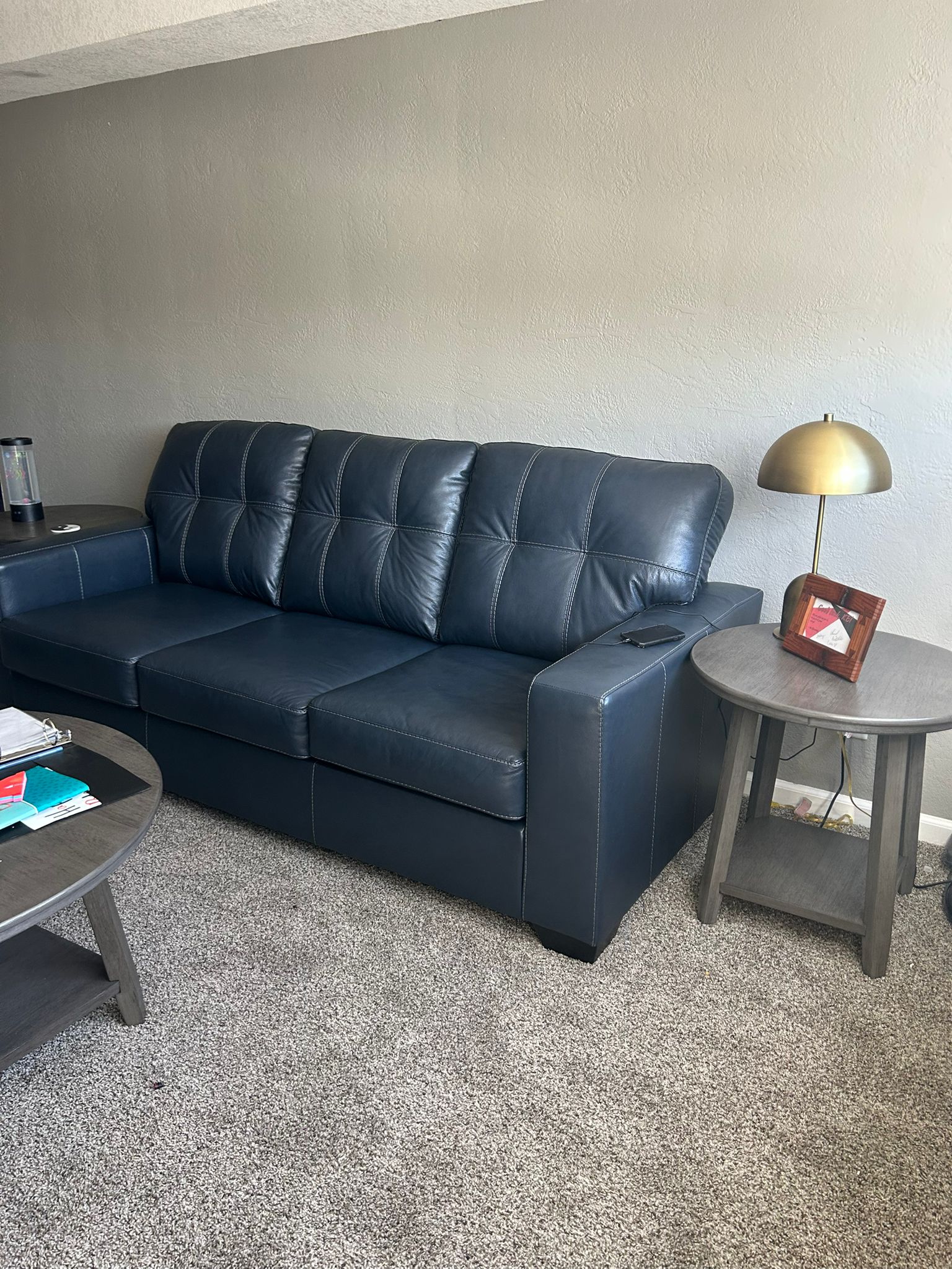 Photo shows a dark leather couch next to a small end table holding a lamp and picture frame.